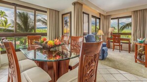 A dining area with a glass table surrounded by wooden chairs, a view of a tropical landscape through large windows, and a balcony with a view of palm trees. - Kaanapali Alii 455 | Hot Tub | Pool | Sunrise Views (Lahaina)
