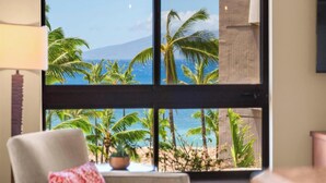 Ein Zimmer mit großem Fenster mit Blick auf einen Strand, Palmen und ein Gebirge.