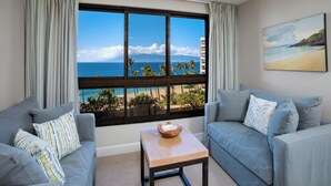 Una habitación con un ventanal que ofrece vistas al océano y a una playa, dos sofás azul claro, una pequeña mesa de madera y un cuadro en la pared.