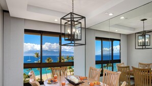 Un comedor con mesa y sillas de madera, ventanas amplias con vista al océano y las montañas, y una lámpara colgante.