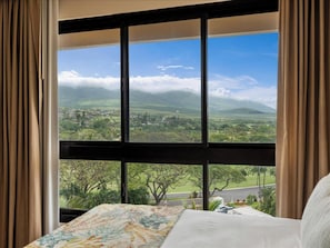 Un dormitorio con un ventanal grande, una cama con cubrecama floral y vistas a un paisaje verde con montañas.