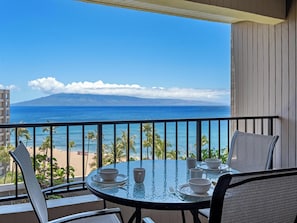 A balcony with a table set for dining, overlooking a beach and ocean with a mountain in the distance.