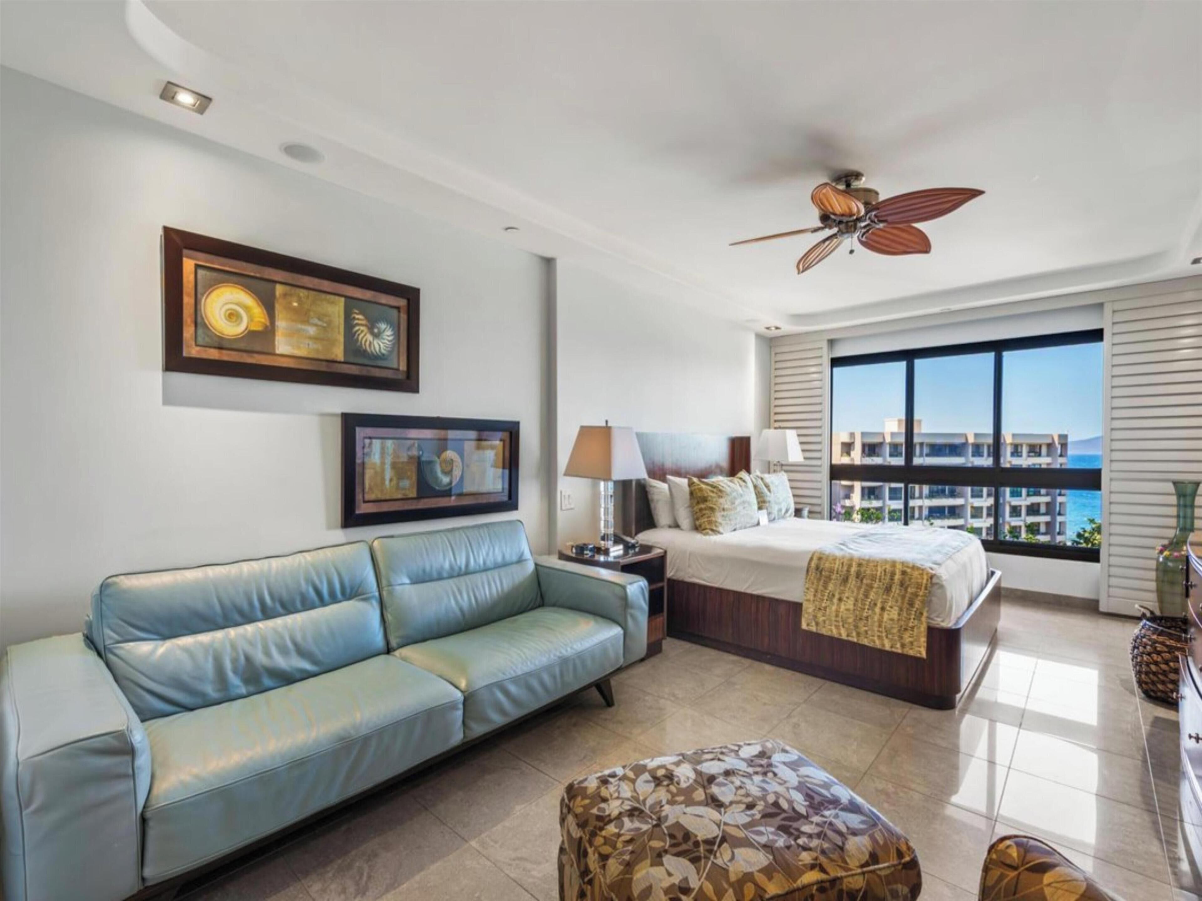 A hotel room with a bed, a blue sofa, a ceiling fan, a large window with a view, and artwork on the wall.