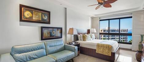 A hotel room with a bed, a blue sofa, a ceiling fan, a large window with a view, and artwork on the wall.