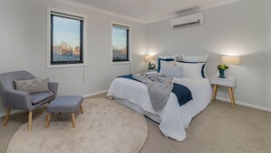 Un dormitorio con cama, una silla, una mesita de noche y dos ventanas.