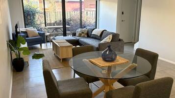 Un salon moderne avec une table en verre, des canapés gris et une grande porte-fenêtre coulissante donnant sur une terrasse.