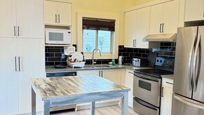 A modern kitchen with light wood cabinets, a stainless steel refrigerator, a built-in oven, a microwave, and a wooden dining table.