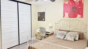 A bedroom with a bed, a desk, a chair, a ceiling fan, and a large window.