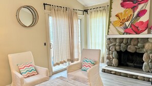 A living room with two white armchairs, a wooden coffee table, a fireplace with a stone surround, and a large floral painting above the fireplace.