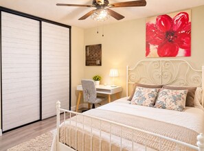 Une chambre à coucher comprenant un lit, un bureau, une chaise, un ventilateur de plafond et un grand tableau représentant des motifs floraux.