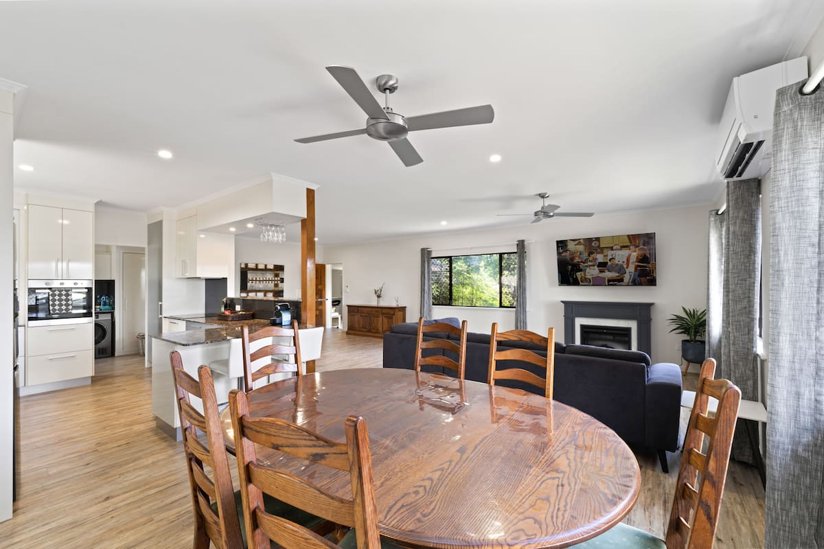 A modern kitchen with white cabinetry, a dining area with a wooden table and chairs, a ceiling fan, and a fireplace.