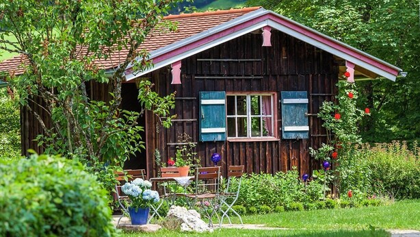 A rustic wooden cabin with a red roof, surrounded by lush greenery and flowering plants. - Vacation apartment Hirscherl, 1-2 persons, 55 sqm (Schönau am Königssee)
