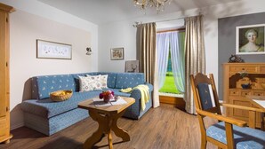 A living room with a blue sofa, wooden table, and a view of a green garden through a window. - Vacation apartment Hirscherl, 1-2 persons, 55 sqm (Schönau am Königssee)