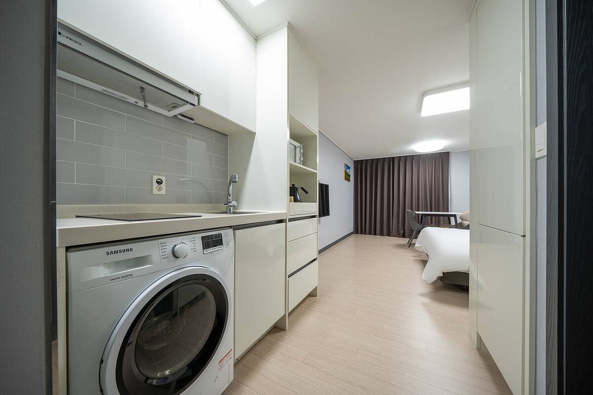 Una habitación de hotel moderna con lavadora, una cama, una mesita pequeña y una silla.