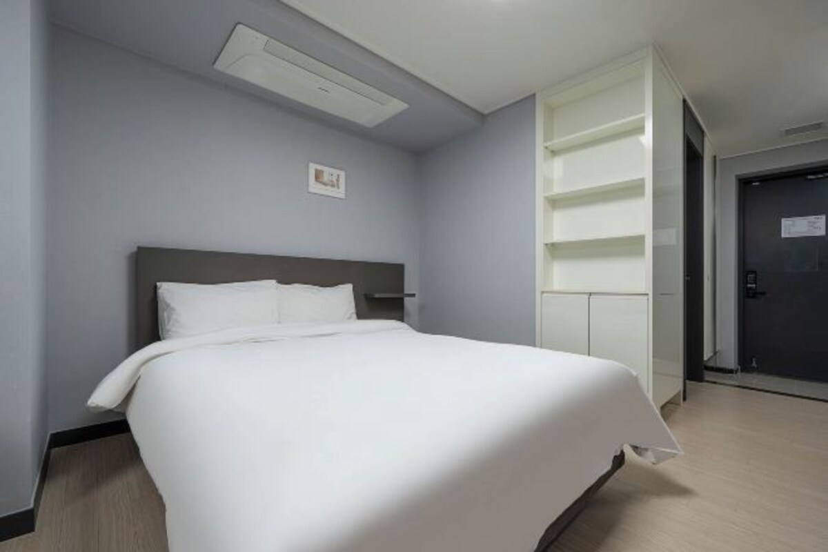 Habitació d'hotel amb un llit, una taula de nit, una prestatgeria penjada a la paret i una porta.