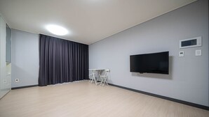 Ein modernes, minimalistisch eingerichtetes Zimmer mit einem Flachbildfernseher, einem kleinen Tisch mit Stühlen und einem großen Fenster mit Vorhängen.
