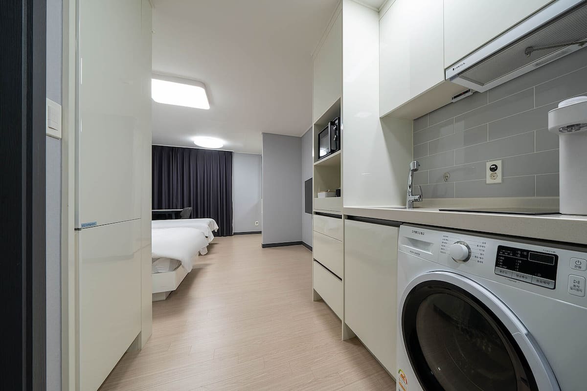 Una habitación de hotel moderna con lavadora, cama, baño con ducha y una pequeña cocina.