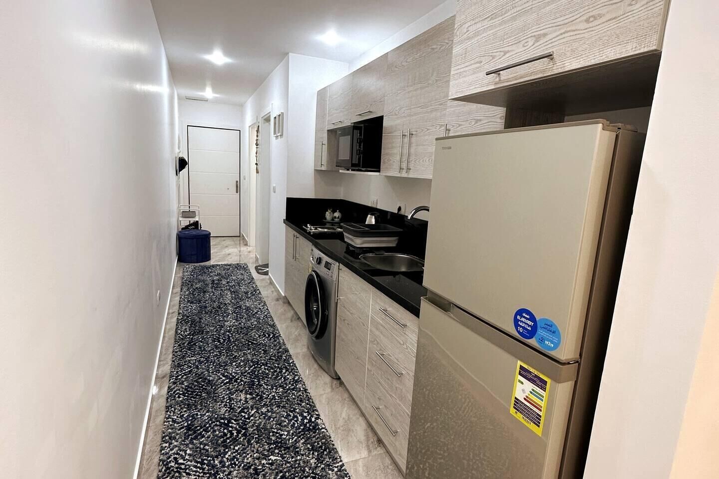 Un pasillo estrecho que da a una cocina con lavadora, refrigerador y microondas.