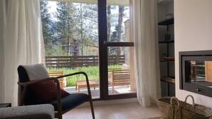 A room with a sliding glass door leading to an outdoor area with a wooden bench and a view of trees and a building. There is a chair with a cushion, a woven basket, and a television on a stand.