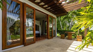 Amplia habitación con grandes puertas correderas de vidrio que dan a un jardín exuberante, techo de madera y cama con colchón blanco.
