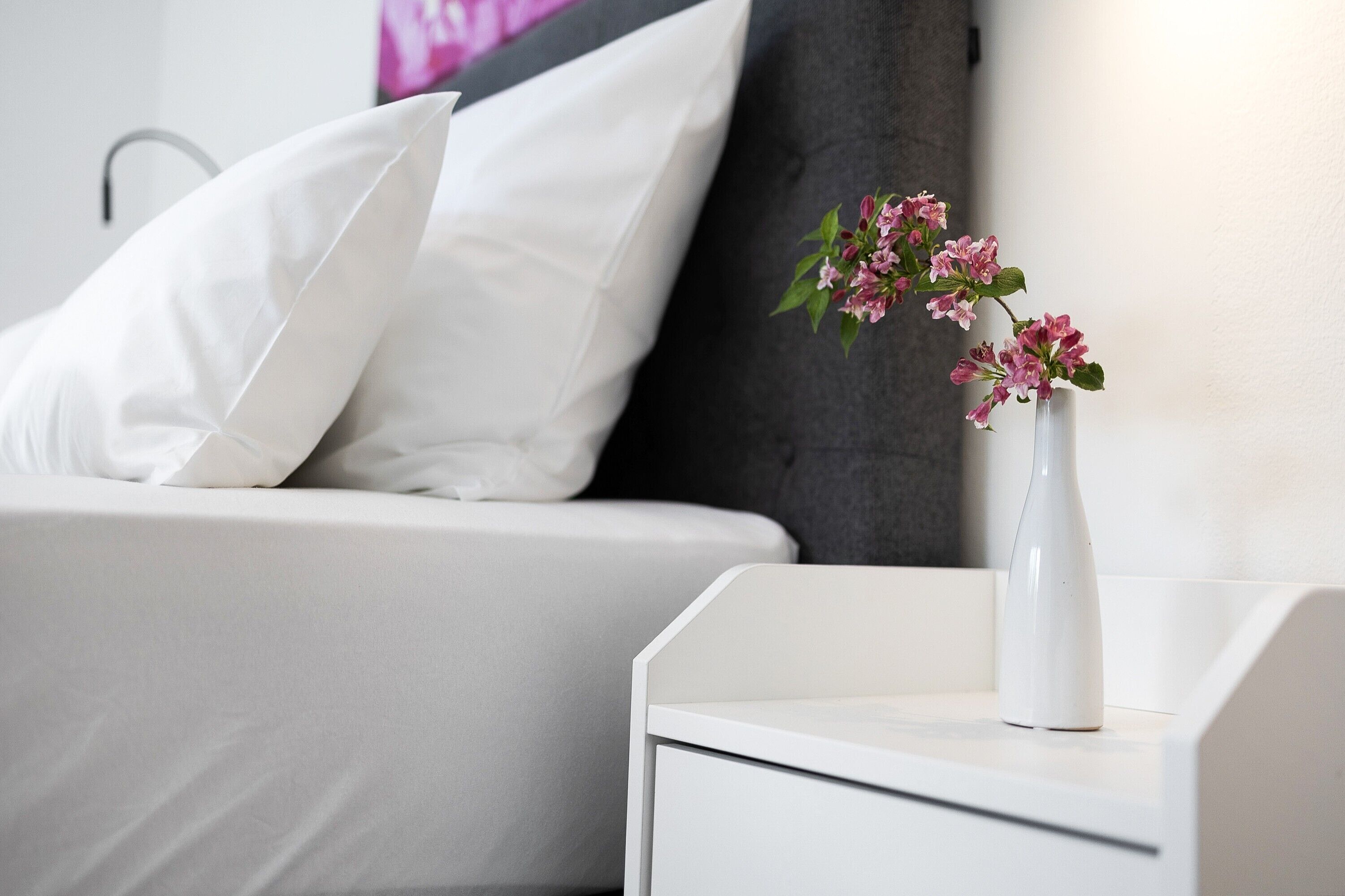 Ein weißer Nachttisch mit einer weißen Vase und rosa Blumen.