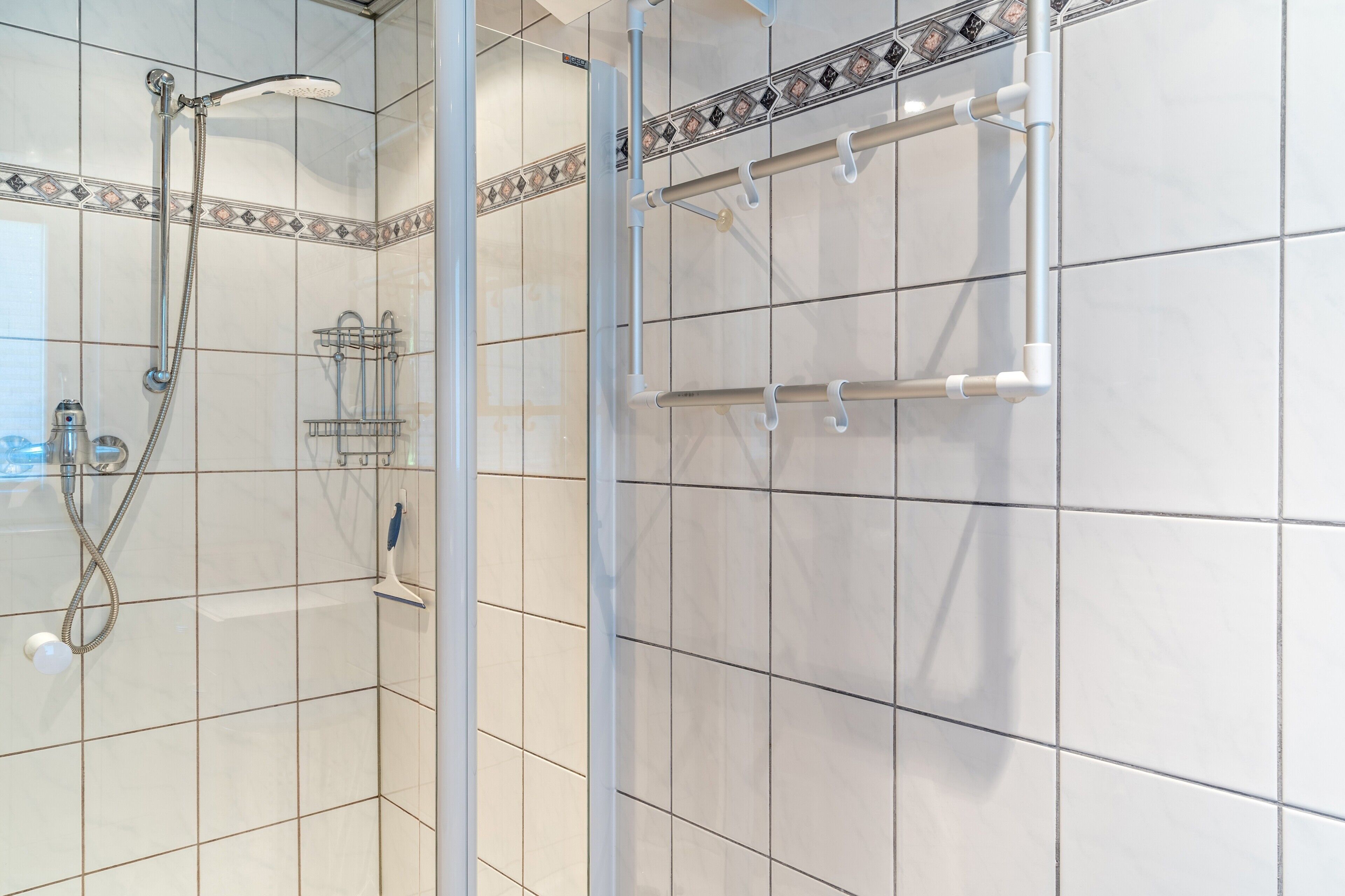 Une salle de bain avec des murs carrelés de carreaux blancs, une douche avec un pommeau de douche amovible, une douche à parois de verre et un porte-serviettes.