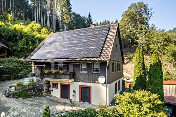 Ein zweistöckiges Haus mit geneigtem Dach, Solaranlagen, einem mit Pflanzen bepflanzten Balkon und einer Steintreppe, die zum Eingang führt.