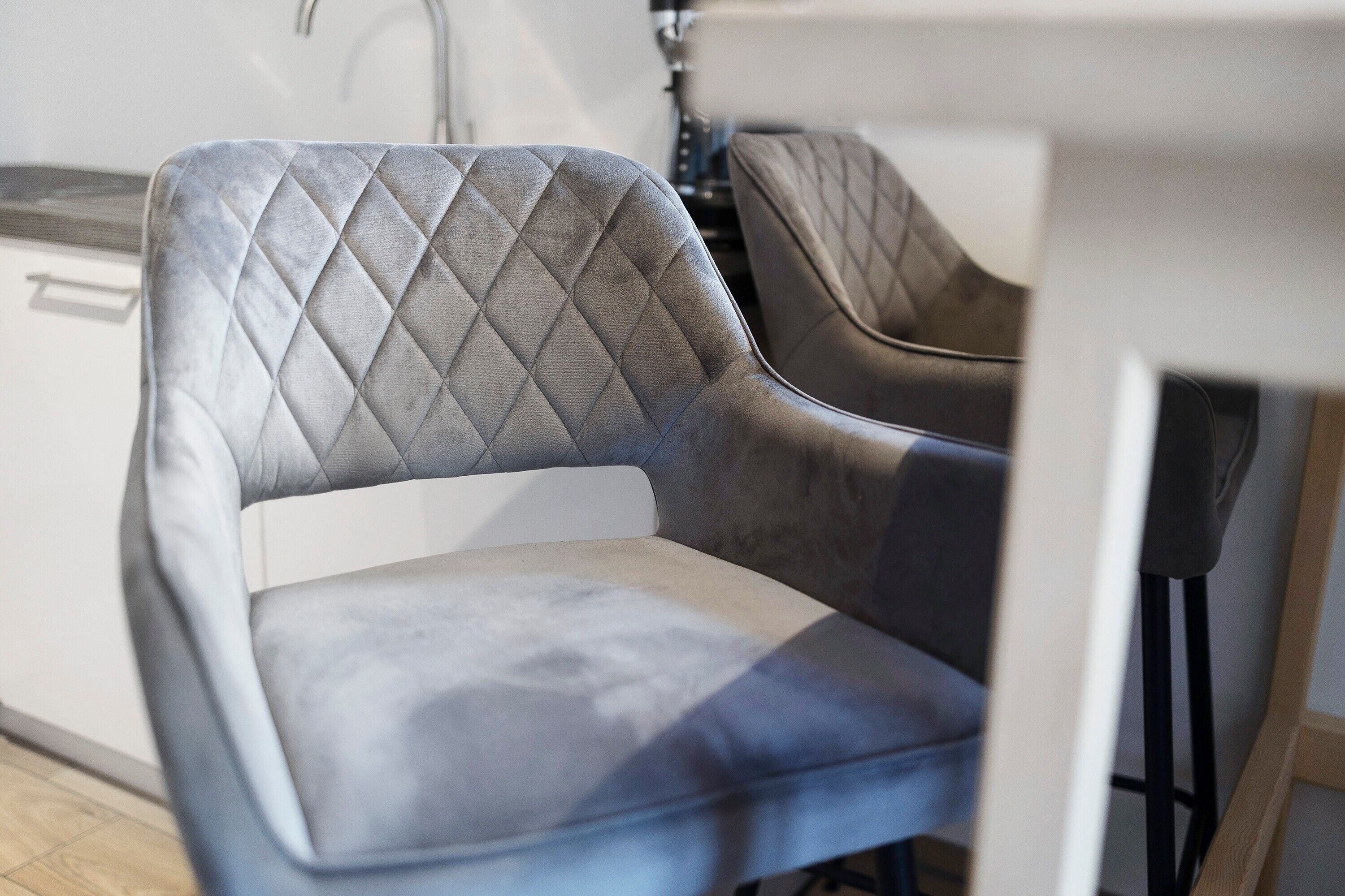 Een moderne keuken met een wit aanrechtblad, een grijze fauteuil met een ruitpatroon rugleuning, en een zwarte stoel met een vergelijkbaar ontwerp.