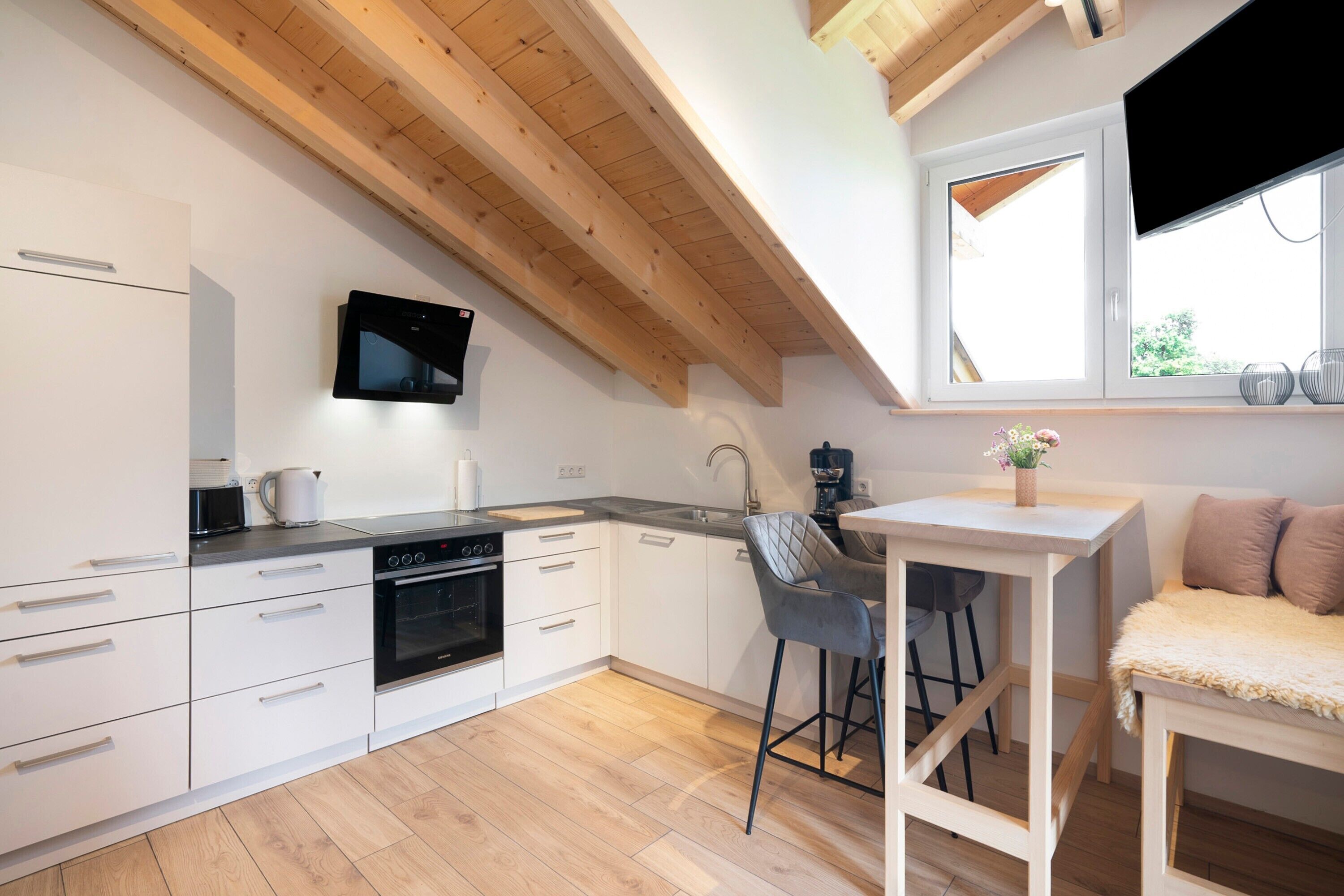 A modern kitchen with white cabinetry, a wooden ceiling, a built-in oven, and a small dining area with a table and chairs.