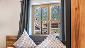 Un dormitorio con cama, almohadones a cuadros, una ventana con vistas a edificios y montañas, y un cabecero de madera.