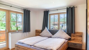 Un dormitorio con una cama de madera, dos ventanas con cortinas y vistas al exterior a zonas verdes, junto a una mesita de noche.