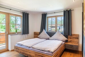 Une chambre avec un lit en bois, deux fenêtres avec des rideaux, une vue sur la verdure et une petite table de chevet.