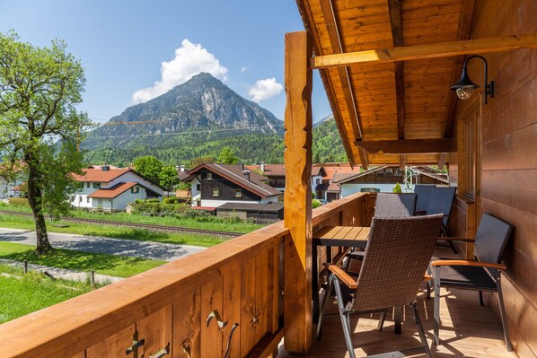 Terraza de madera con sillas de mimbre y una mesa, con vistas a una montaña y casas.