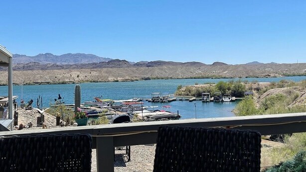 Vista del lago con barcos amarrados, montañas al fondo y un área de patio con sillas.