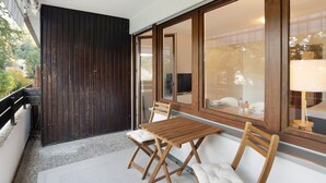 Un balcon avec une table et des chaises en bois, une porte en bois foncé et de grandes fenêtres donnant sur l’extérieur.