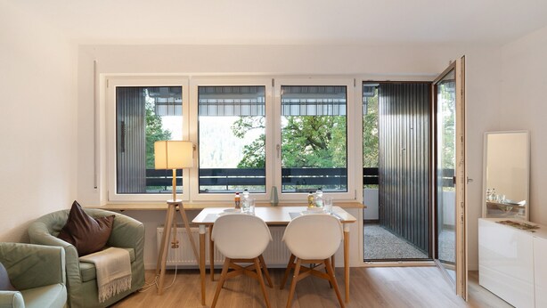 Un salon moderne comprenant un canapé, une table à manger avec des chaises, une lampe et de grandes fenêtres donnant sur un paysage verdoyant.