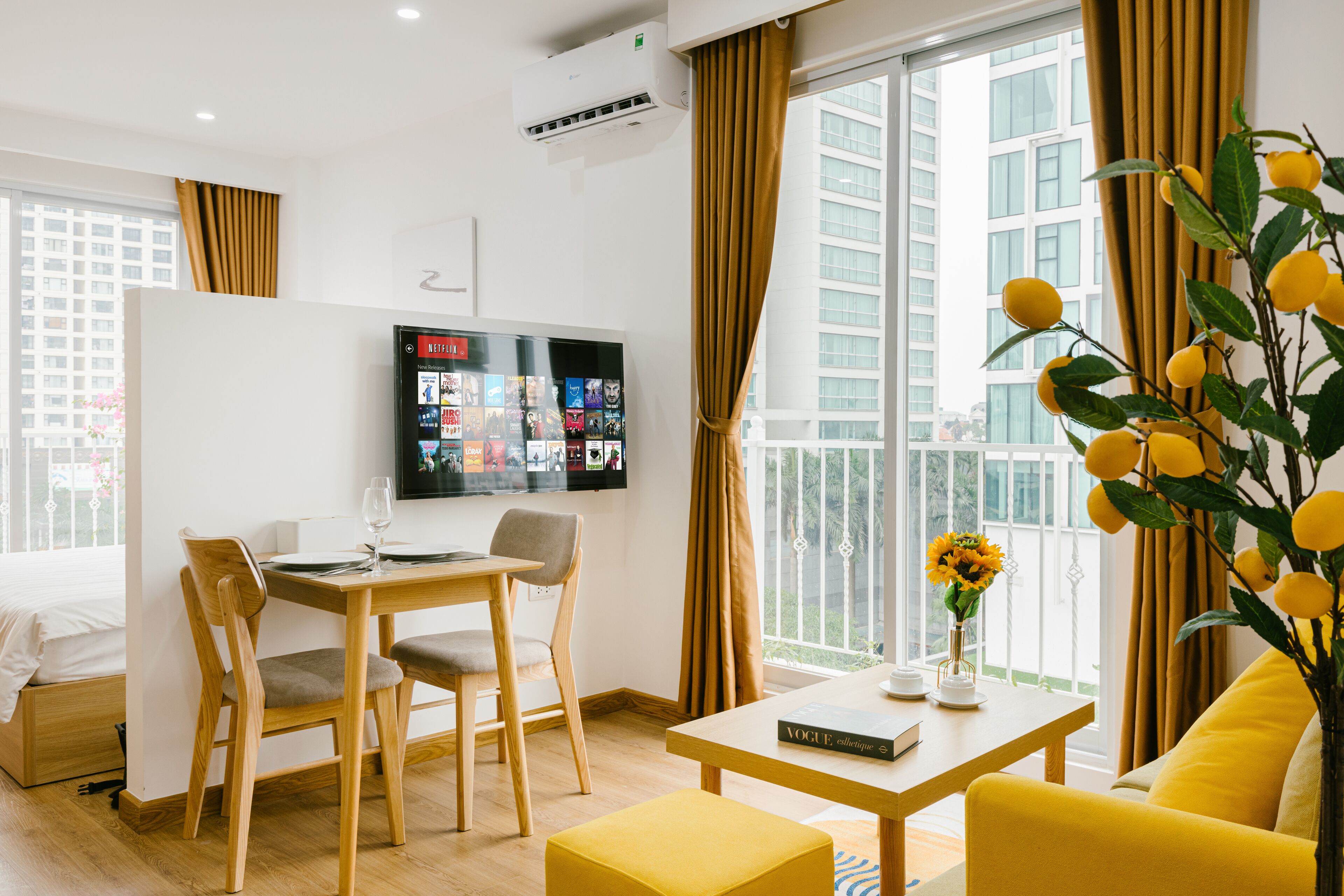 A modern hotel room with a dining area, a sofa, a TV mounted on the wall, and a large window with curtains.