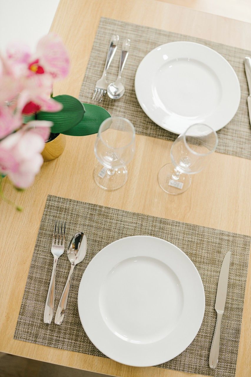 A table set with white plates, cutlery, and wine glasses, with a vase of flowers on the left.