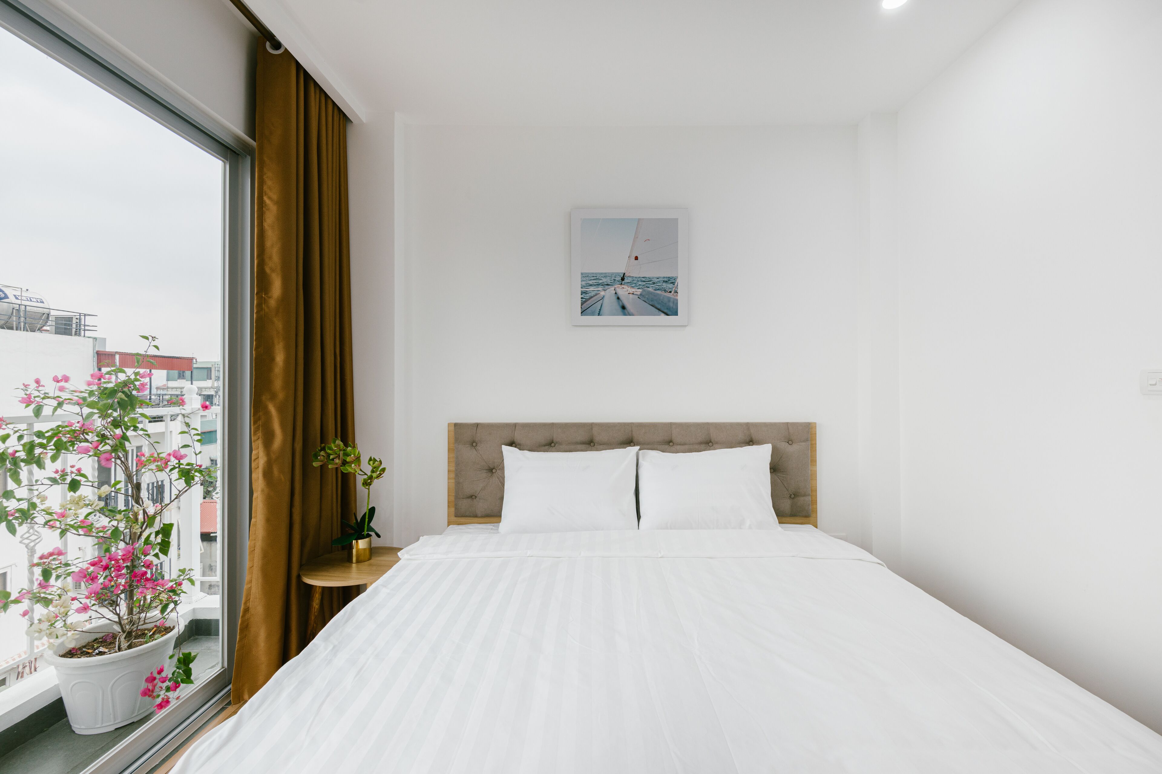 Une chambre d’hôtel avec un grand lit, une fenêtre donnant sur des immeubles, une plante en pot et un tableau encadré au mur.