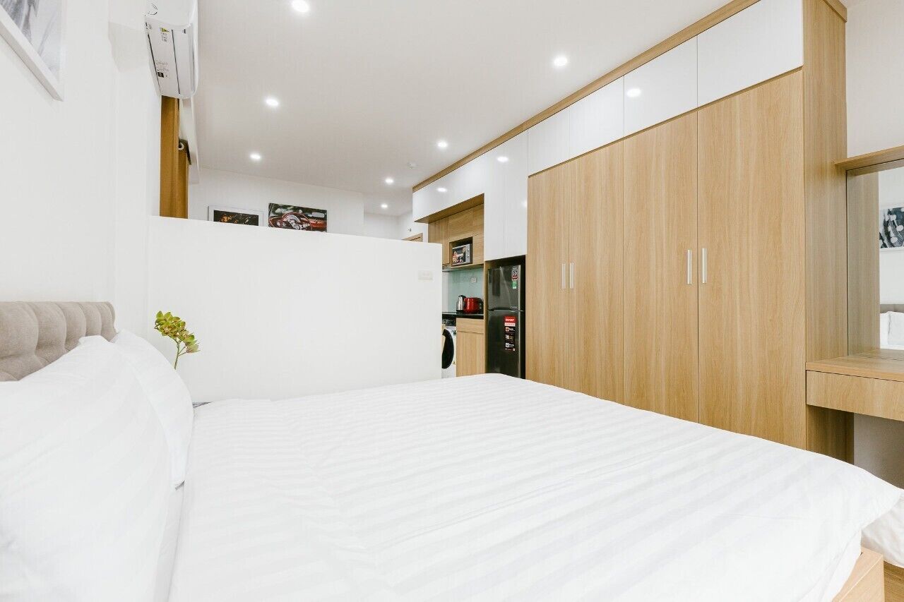 A modern bedroom with a large bed, a wooden wardrobe, and a kitchenette in the background.