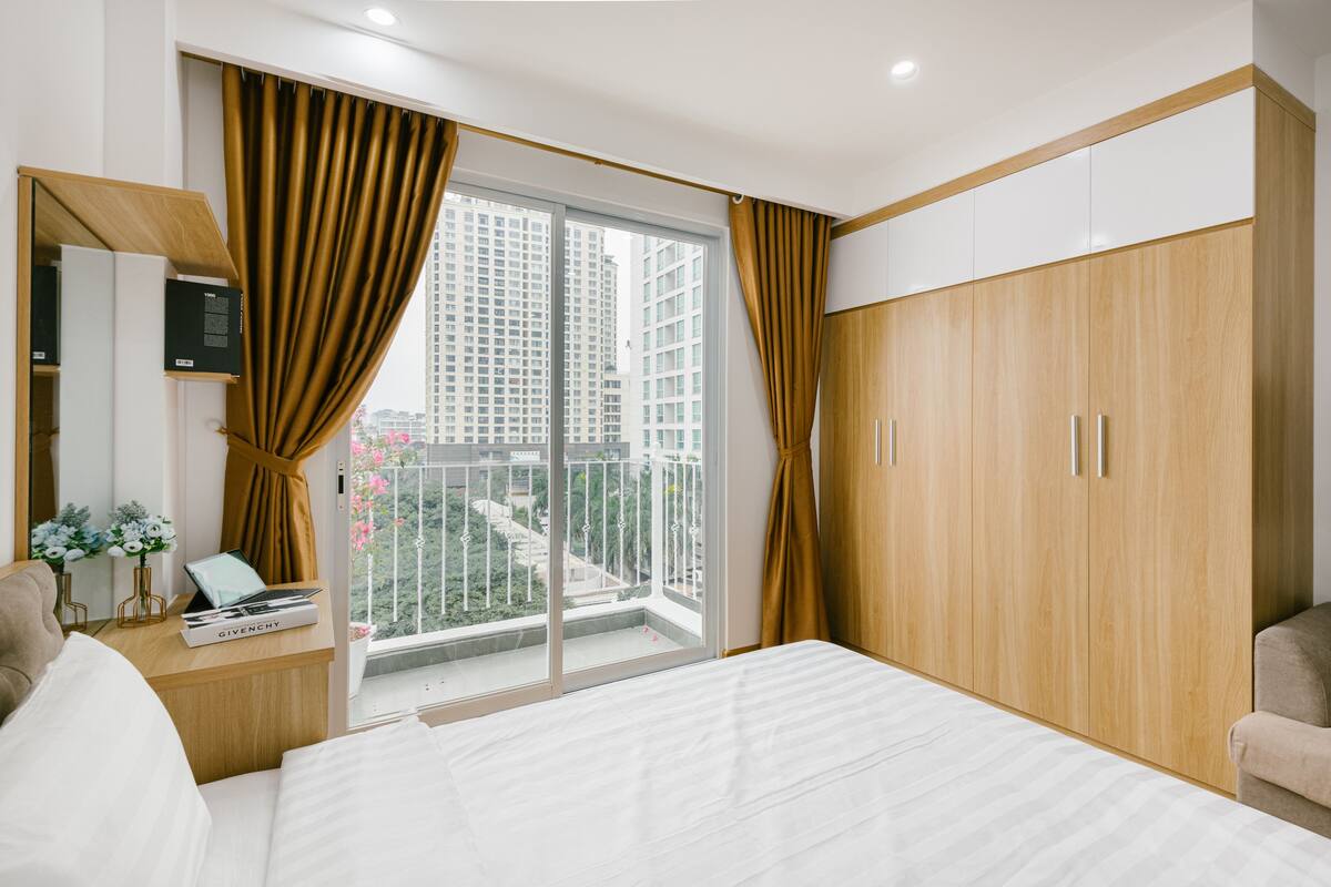 Ein modernes Hotelzimmer mit einem großen Bett, einem Holzschrank, einem Schreibtisch mit einem Laptop und einem Balkon mit Blick auf die Stadt.