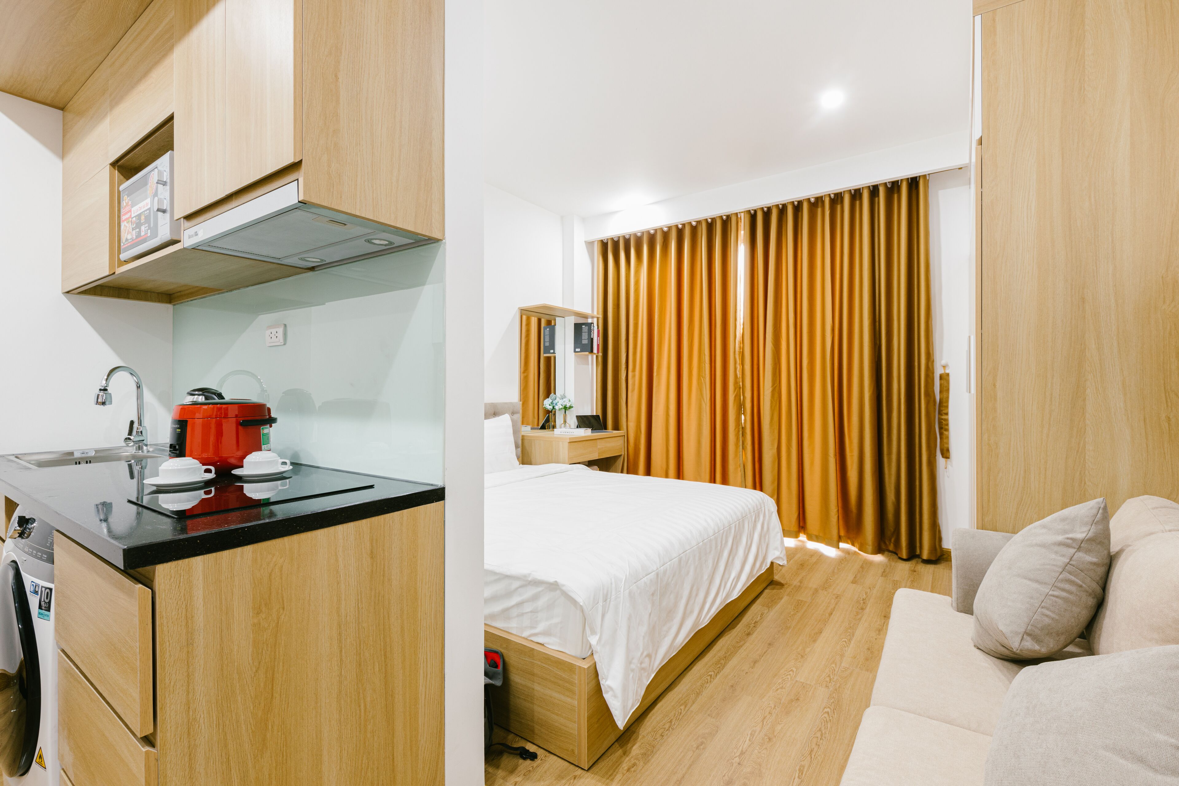 A hotel room with a kitchenette, a bed, a sofa, and a large window with curtains.
