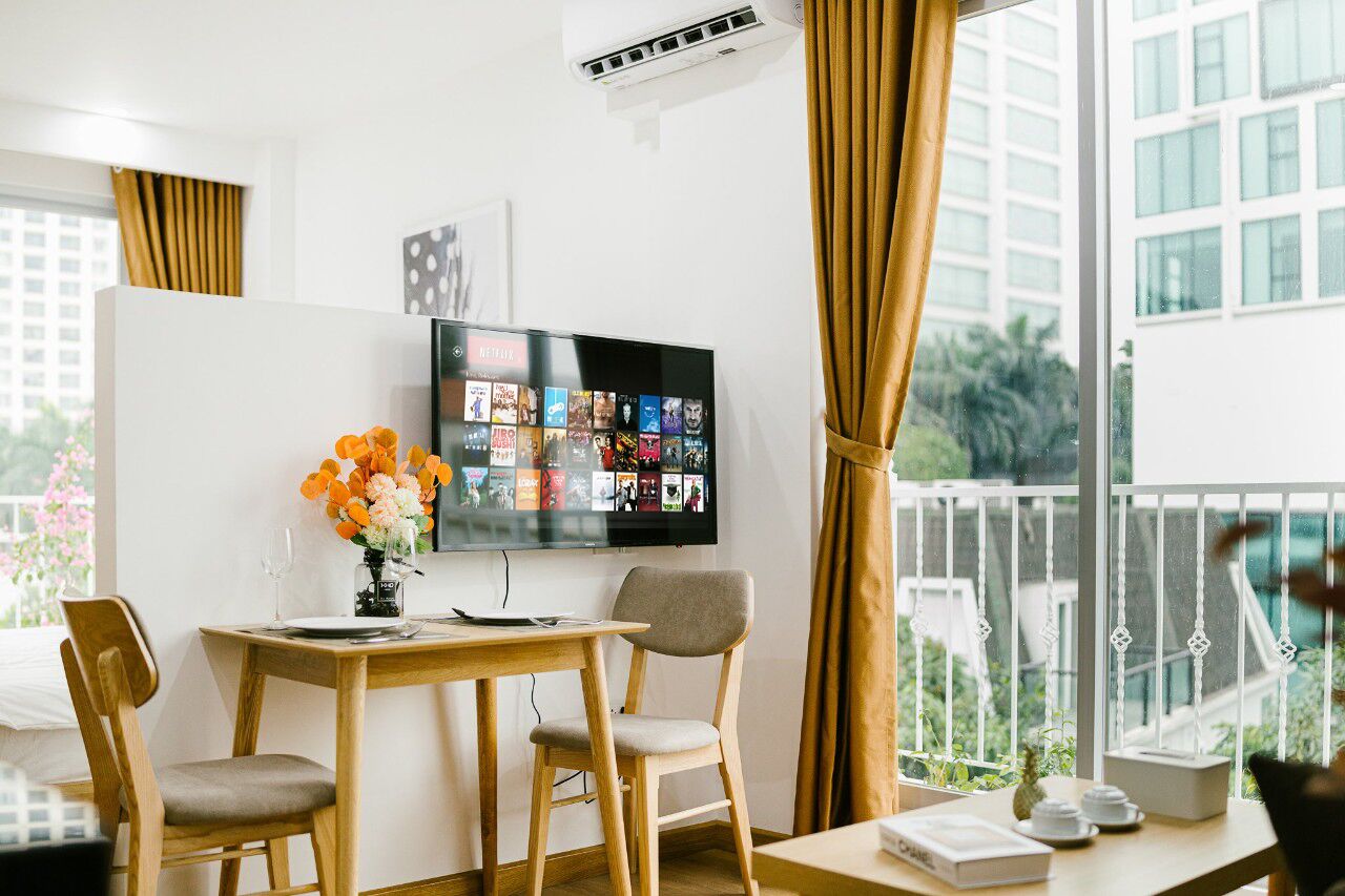 A modern living room with a flat-screen TV mounted on a white wall, a wooden dining table with two chairs, a vase of flowers, and a balcony with a view of buildings and greenery.