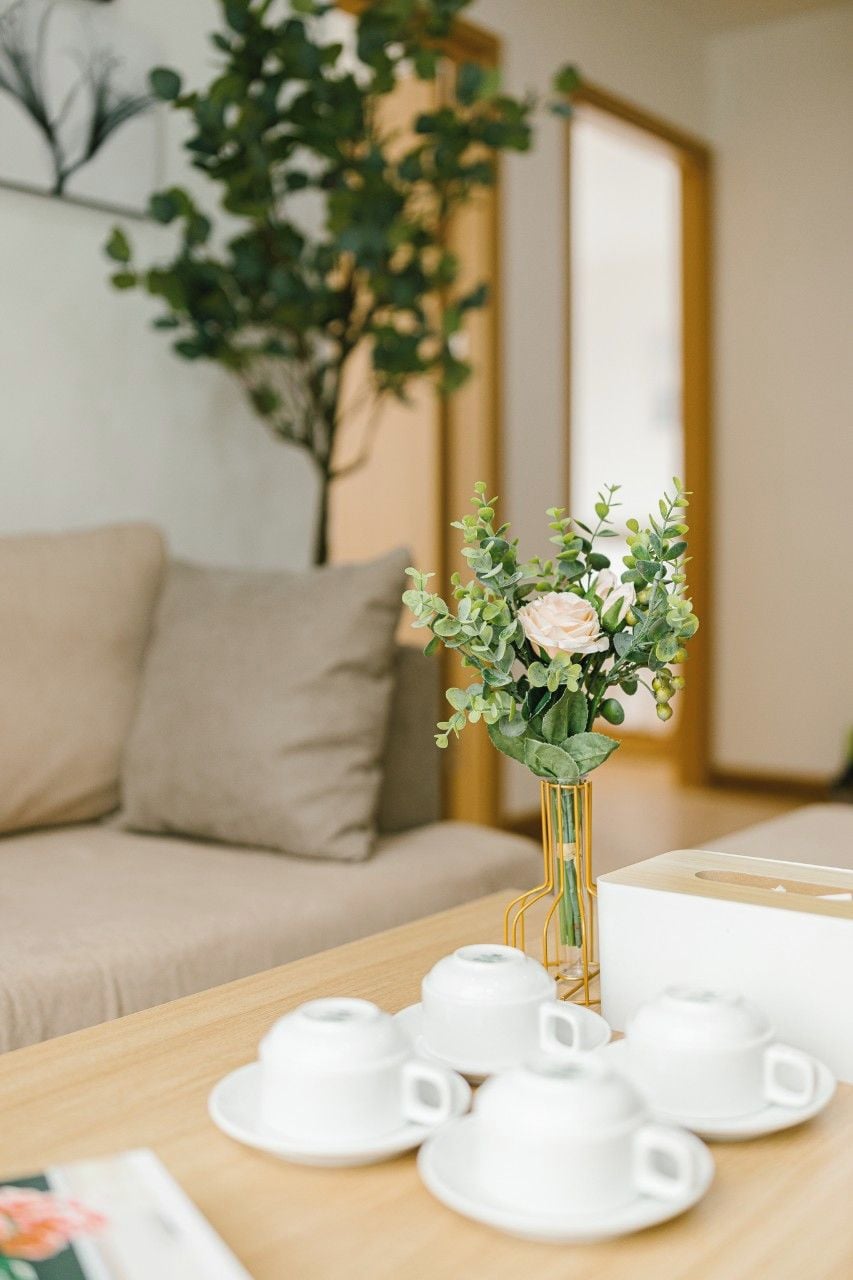 Ein Zimmer mit einem beigefarbenen Sofa, einem Holztisch mit weißen Teetassen und Untertassen sowie einer Vase mit Grünpflanzen und einer einzelnen Blume.