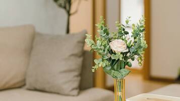 Ein Zimmer mit einem beigefarbenen Sofa, einem Holztisch mit weißen Teetassen und Untertassen sowie einer Vase mit Grünpflanzen und einer einzelnen Blume.