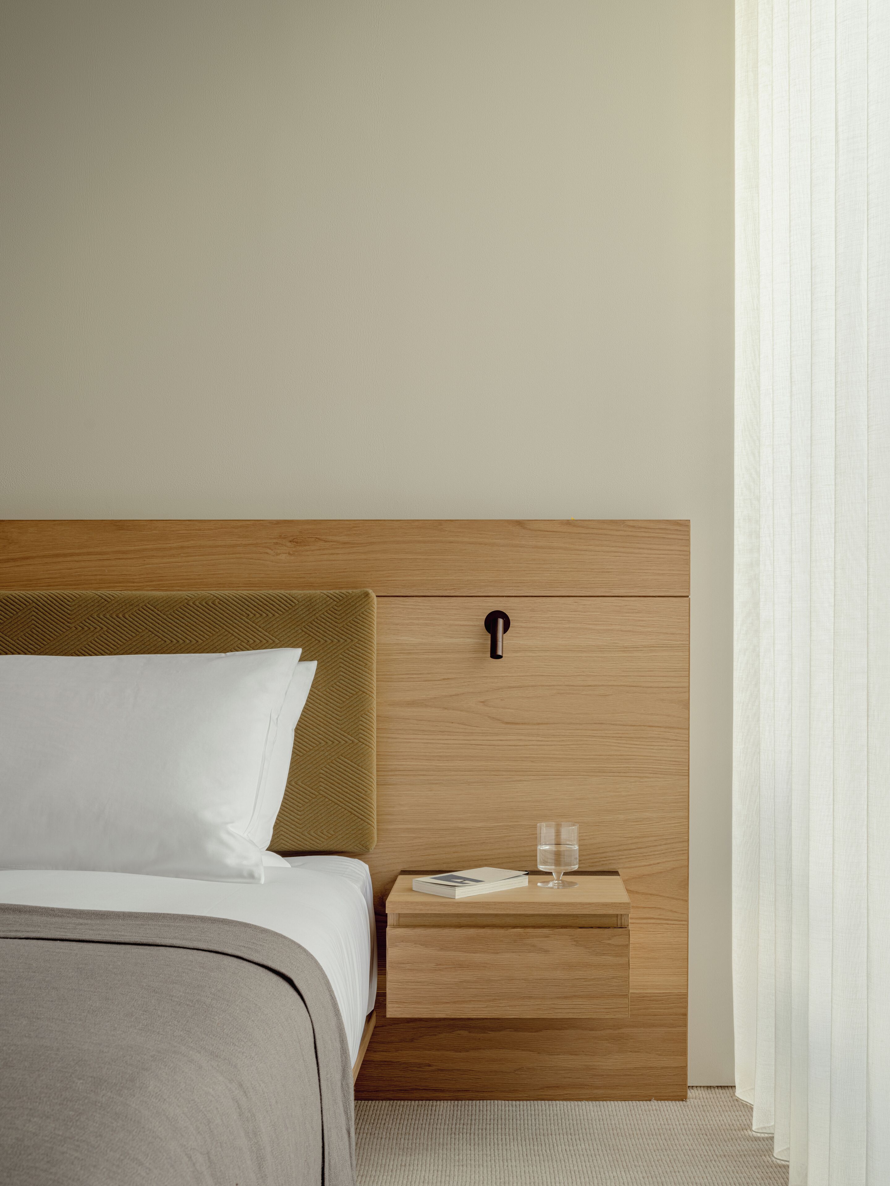 Cama bem arrumada com cabeceira de madeira, mesa de cabeceira com um copo de água e uma gavetinha.