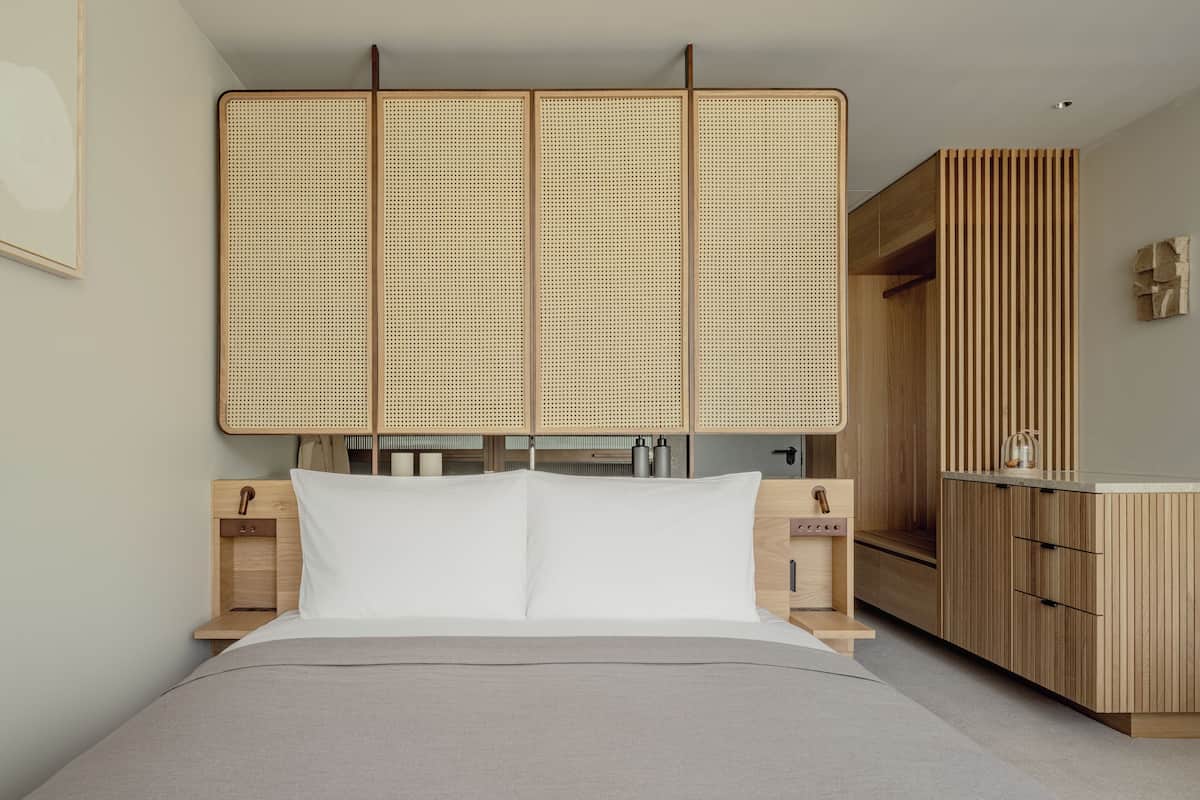 A modern hotel room with a bed, wooden headboard, bedside tables, and a kitchenette area.