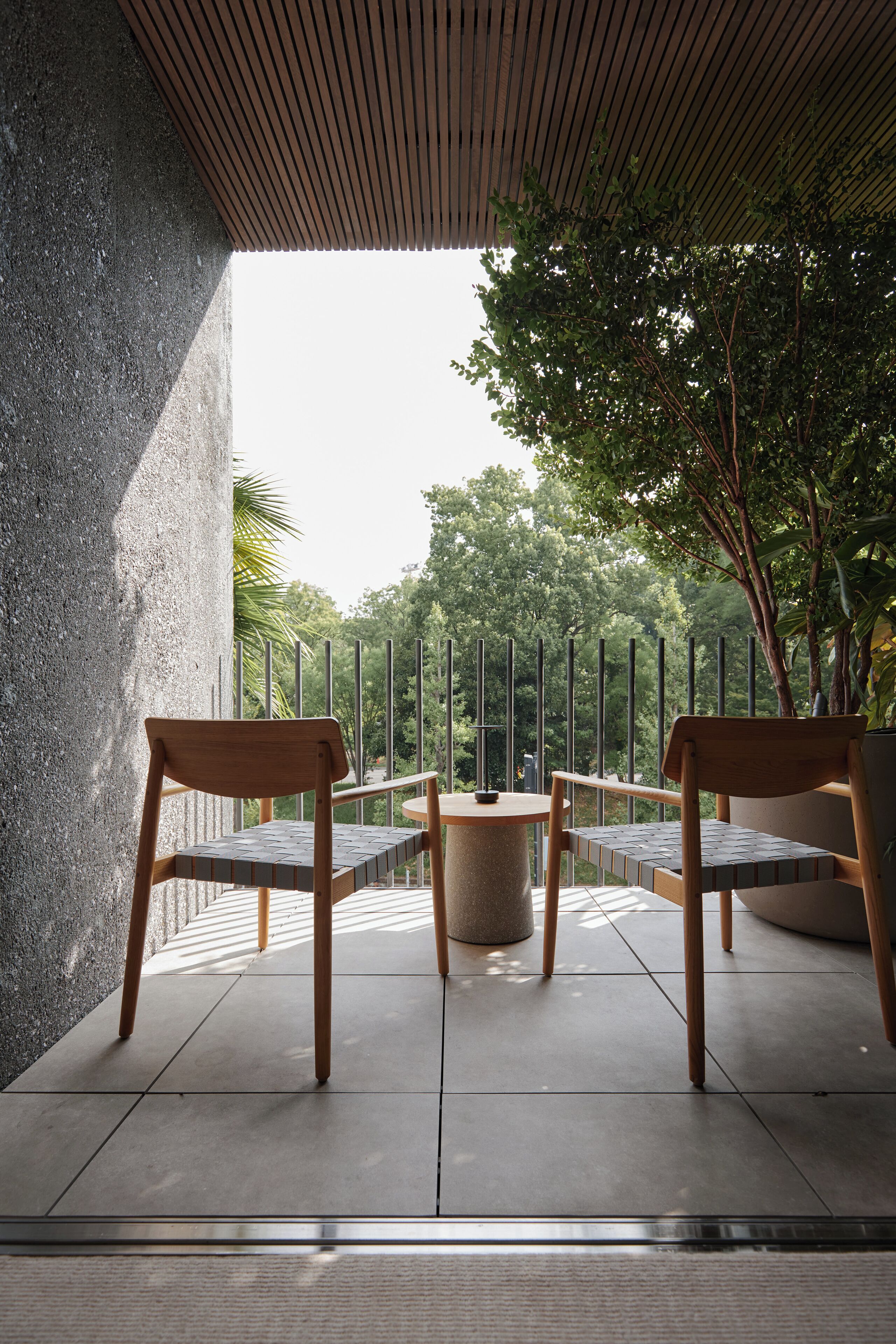 A modern outdoor seating area with two wooden chairs and a round table, surrounded by a concrete wall and a metal fence, overlooking a lush green park.