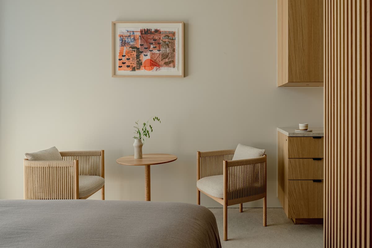 A room with two wicker chairs, a small round table with a vase, a bed with a grey blanket, a wooden cabinet, and a framed artwork on the wall.