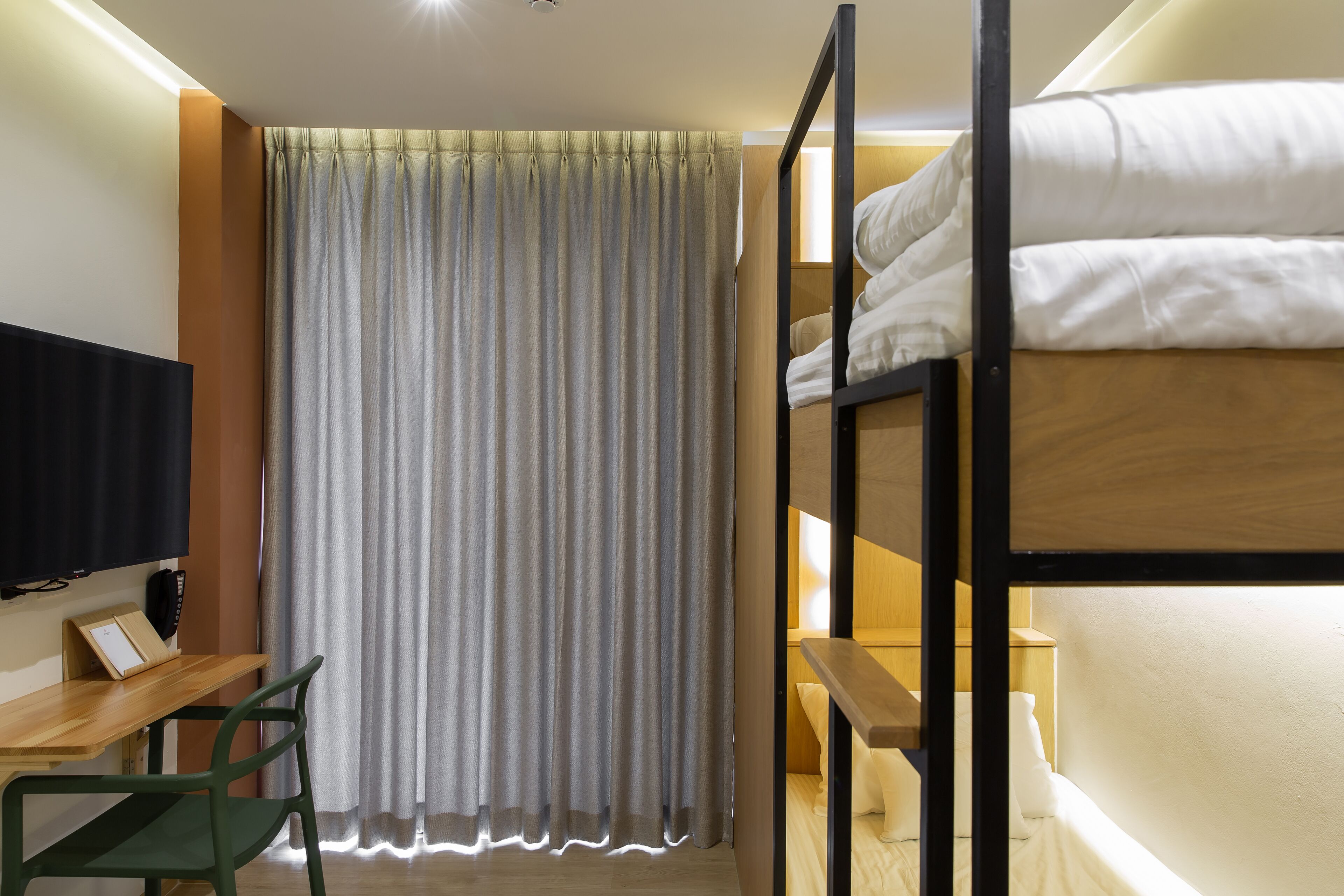A bunk bed with white bedding, a desk with a chair, a television, and a curtain.